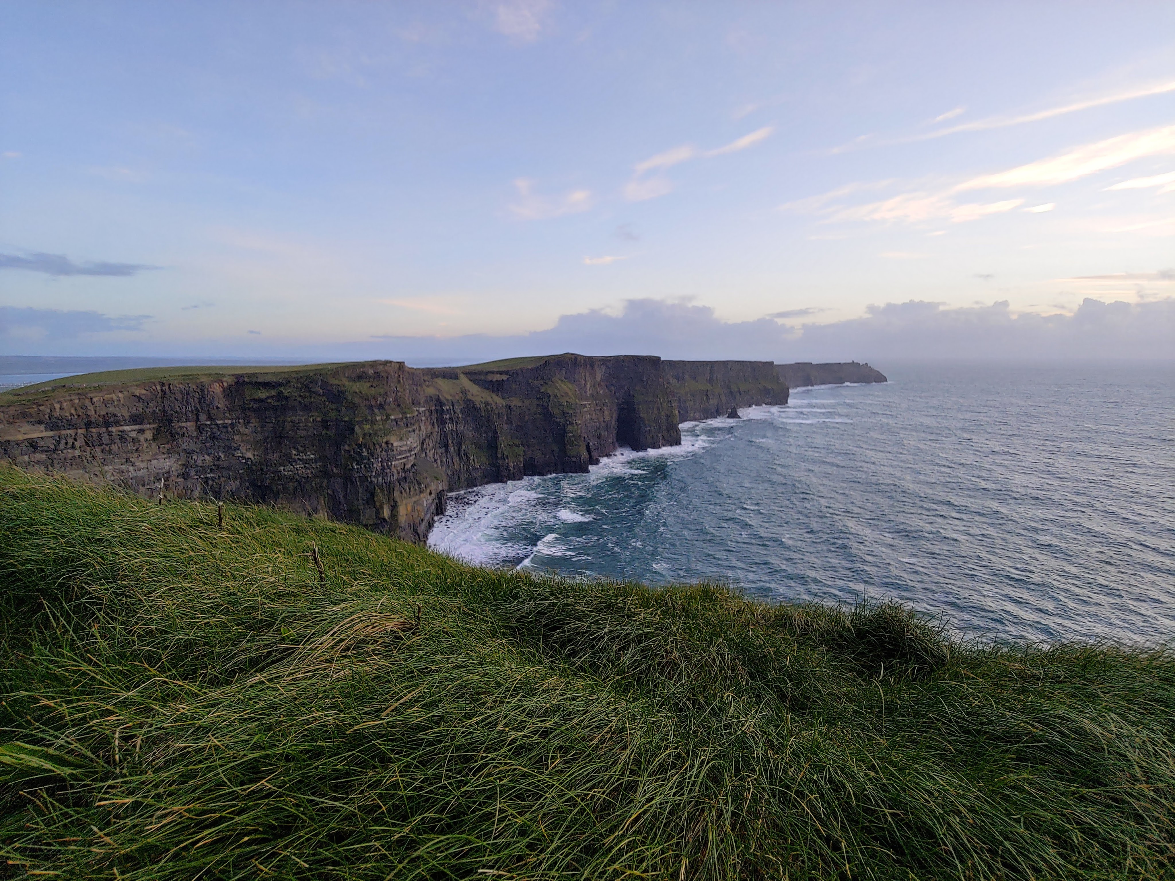 Panoramic view of the Cliffs of Moher in Ireland, with lush green grass overlooking dramatic coastal cliffs and the Atlantic Ocean beneath a pastel evening sky.