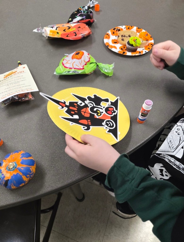 Halloween craft table with a yellow paper-plate haunted-house decoration, glue stick, painted pumpkin, eyeball treat, and pumpkin-themed plate of cupcakes and a cookie.
