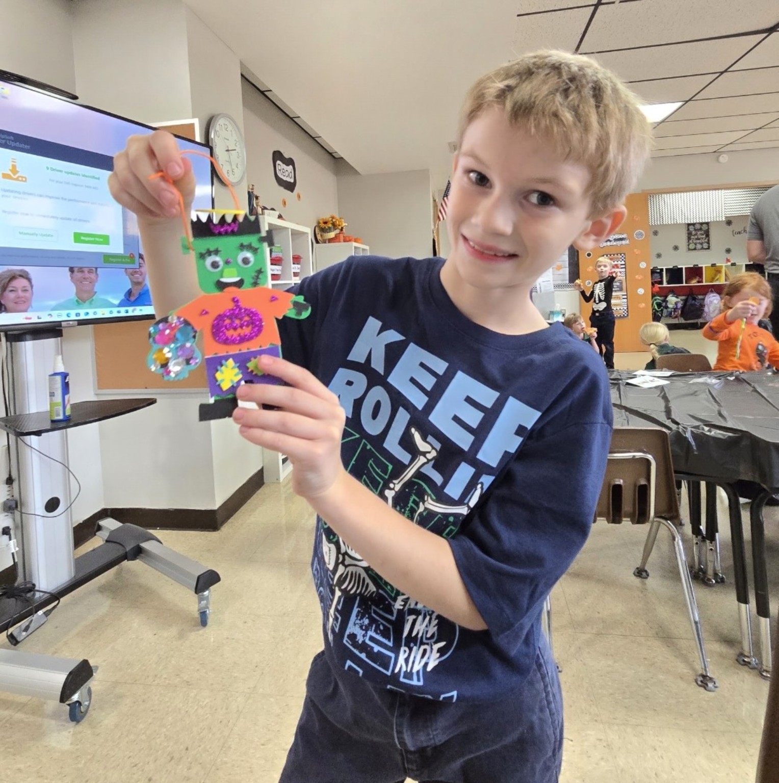 Young boy proudly displaying a colorful handmade Frankenstein Halloween decoration with glitter and paper cutouts during a children’s craft activity.