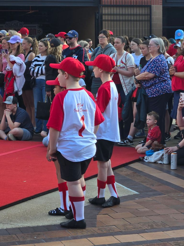 Young Irish dancers in red and white O'Faoláin Academy uniforms stand near a performance area surrounded by spectators.
