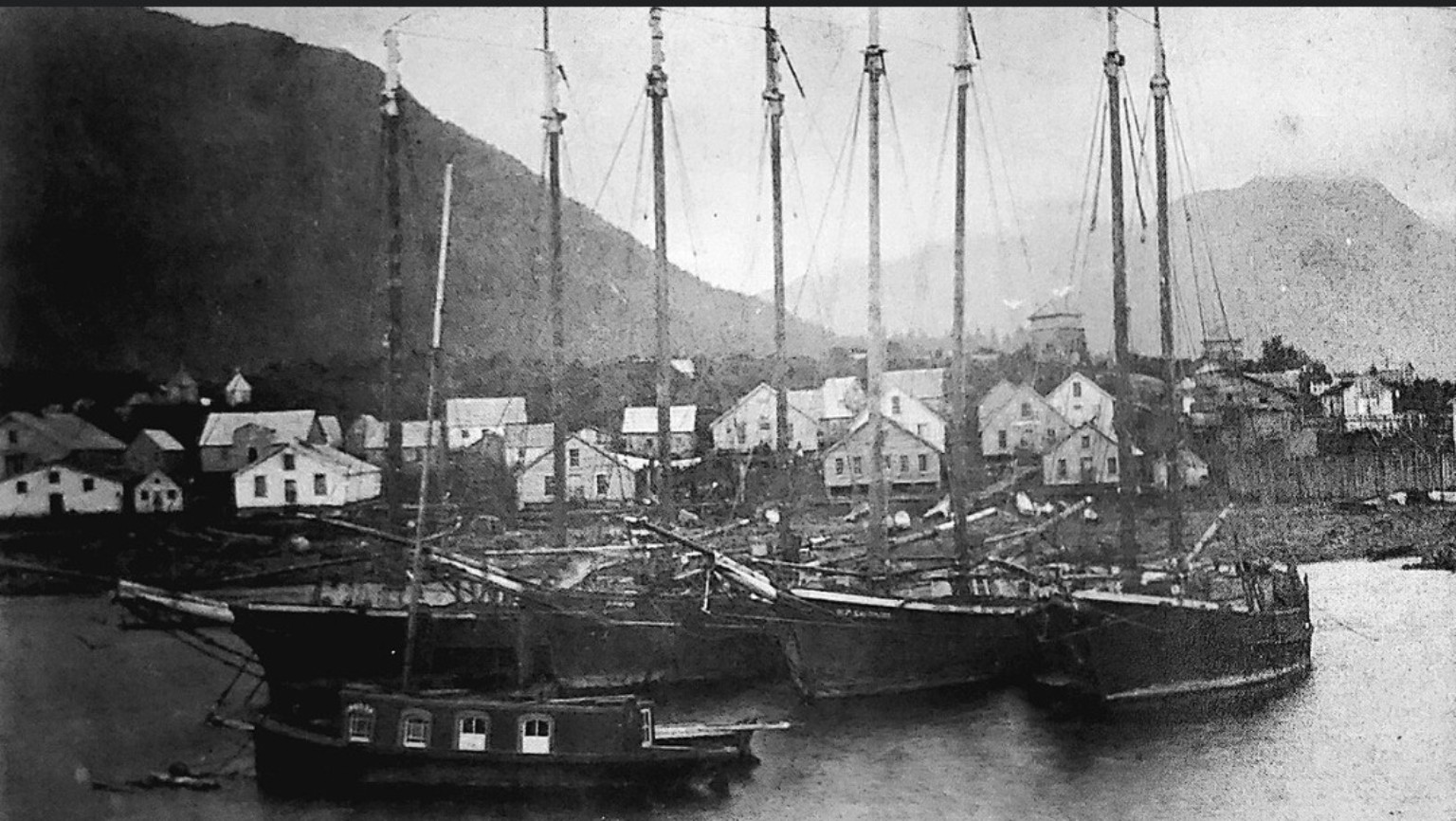 Historic black-and-white photograph of Sitka, Alaska, circa 1886, with tall-masted sailing ships, a Tlingit village along the waterfront, and mountains behind.