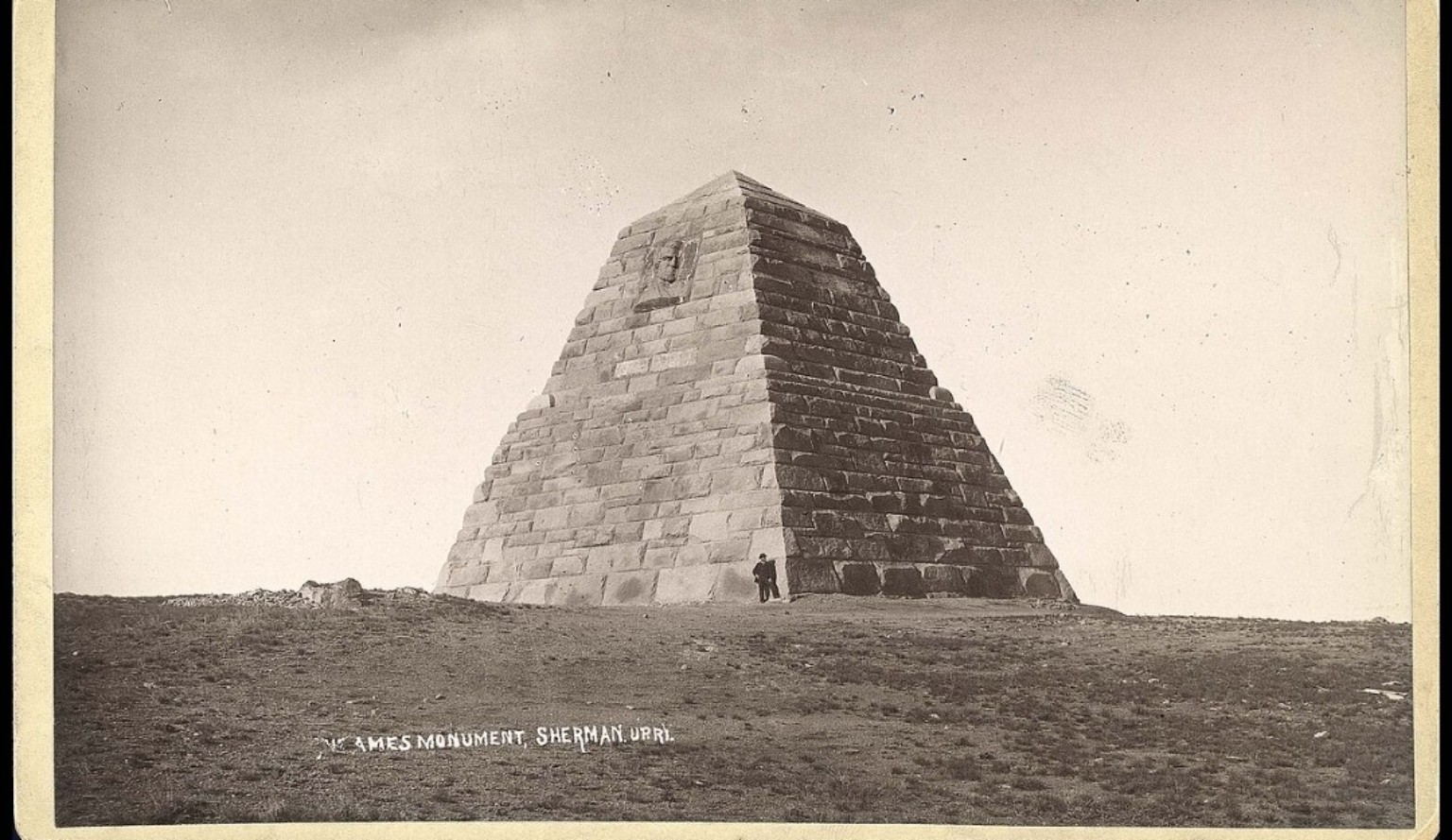 Historic sepia photograph of the massive stone Ames Monument pyramid near Laramie, Wyoming, with a person standing at its base for scale.