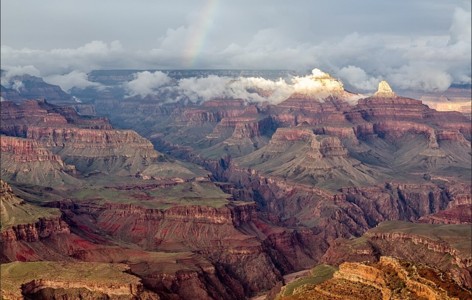 Layered red and purple cliffs of the Grand Canyon seen from Hopi Point beneath dramatic clouds, sunlight and a faint rainbow.