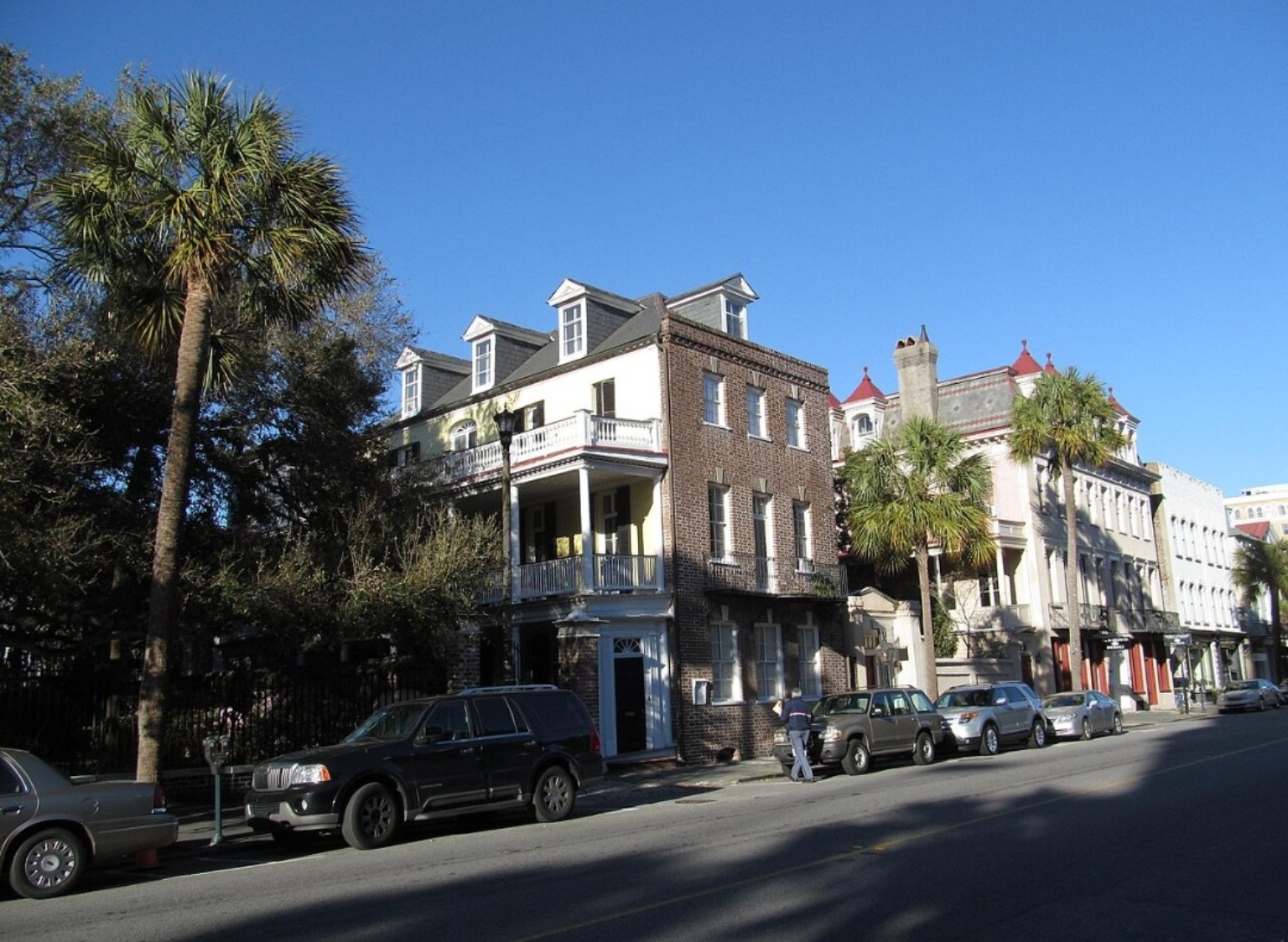 Historic brick and stucco buildings with balconies and dormer windows along a Charleston street lined with palm trees.