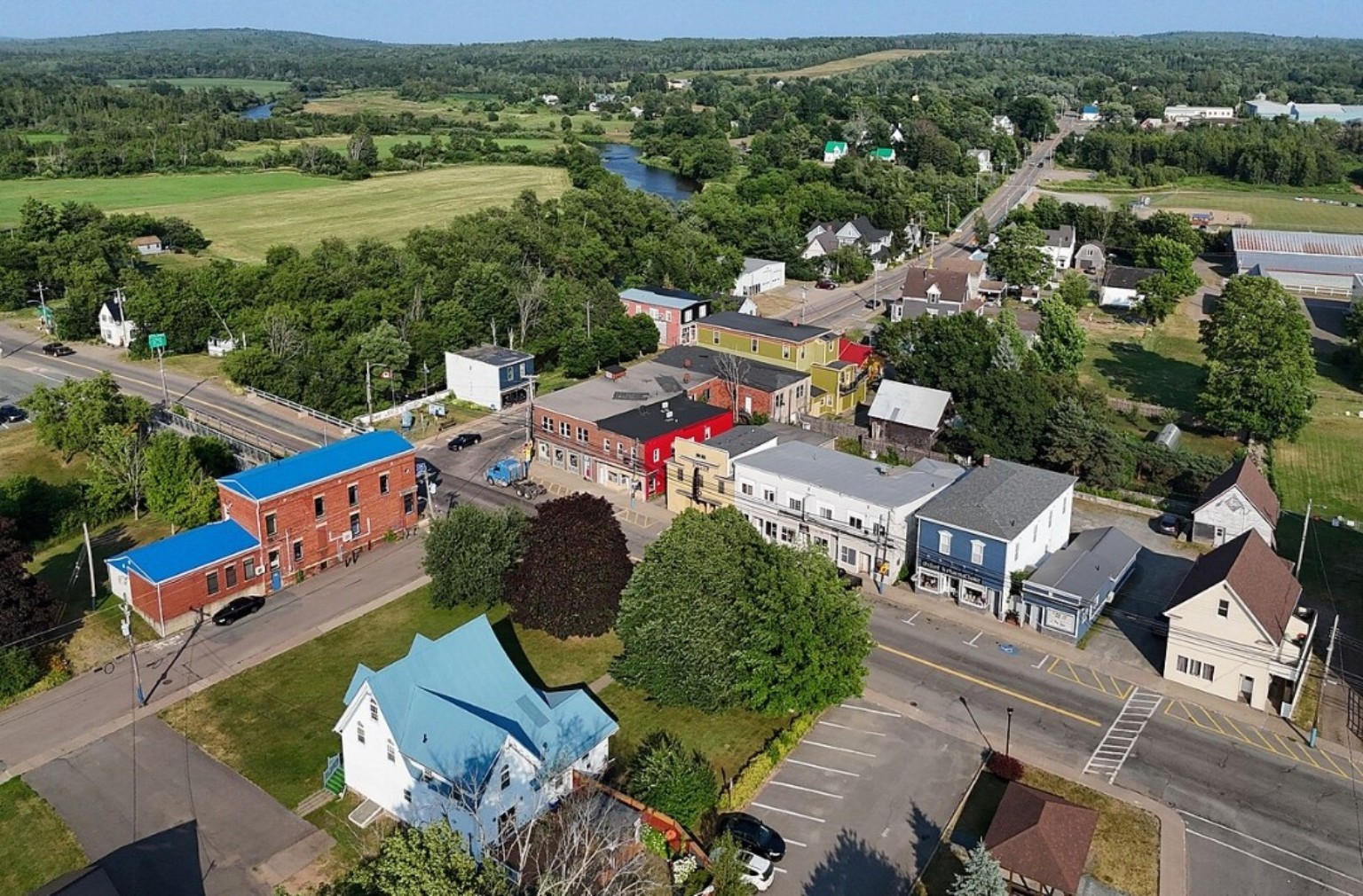 Aerial view of Oxford, Nova Scotia, showing colorful storefronts, a river, tree-lined streets, and surrounding green countryside.