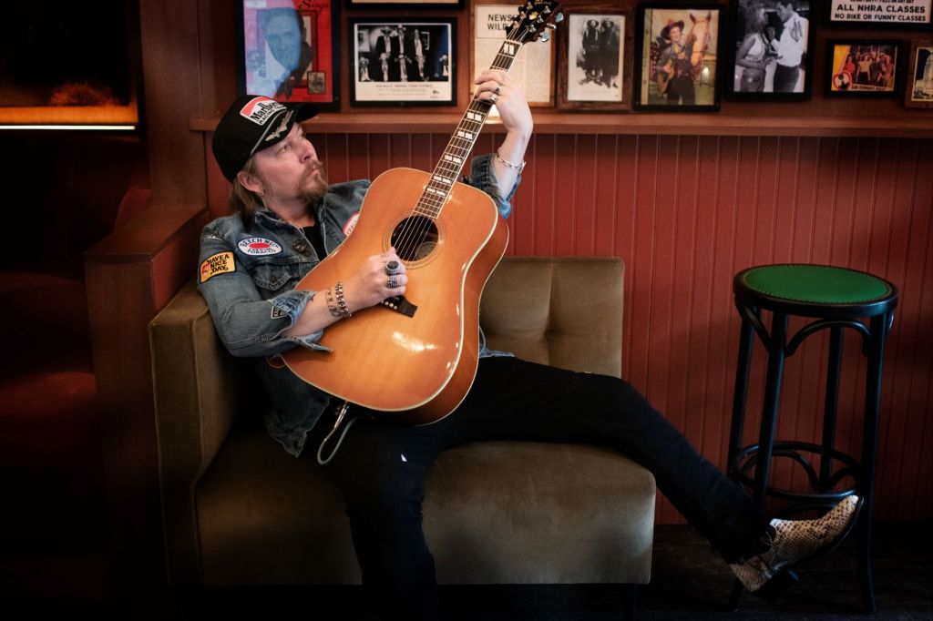Drew Ryder Smith playfully raising an acoustic guitar while seated indoors, photographed by Kim Mancuso.