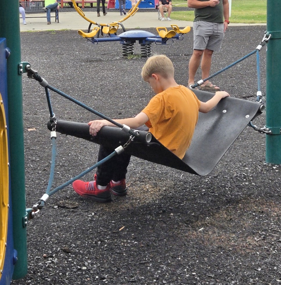 Boy in an orange shirt sitting alone on a suspended playground seat, looking down while other children play in the background.